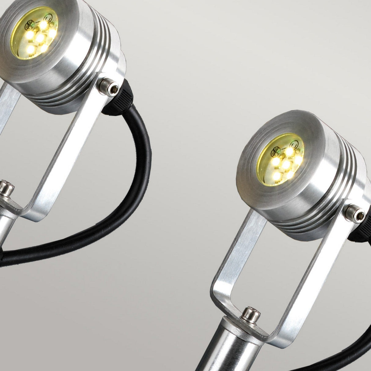 Close-up of two Spennymoor Kit spotlights featuring silver metal housings and flexible black cords, ideal for 12V garden illumination. Set against a light gray background, these cylindrical lights highlight their bright yellow bulbs, offering versatile outdoor lighting solutions.