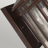 A close-up of the Chedworth 1 Light Outdoor Wall Lantern showcases its durable cast aluminum construction with a bronze finish. The rectangular fixture is enhanced by textured glass panels and is tastefully mounted on a matching bronze wall plate, set against a light gray background.