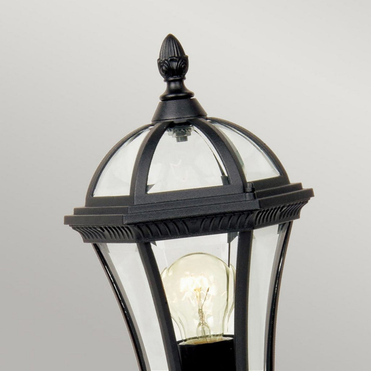 The Ledbury 1 Light Pedestal Lantern - Black is an outdoor lantern crafted from cast aluminum, featuring a classic black finish and a clear glass enclosure that showcases an exposed bulb. Its top is adorned with a decorative finial, set against a soft, neutral gray background for added elegance in your exterior lighting decor.