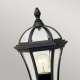 The Ledbury 1 Light Pedestal Lantern - Black is an outdoor lantern crafted from cast aluminum, featuring a classic black finish and a clear glass enclosure that showcases an exposed bulb. Its top is adorned with a decorative finial, set against a soft, neutral gray background for added elegance in your exterior lighting decor.