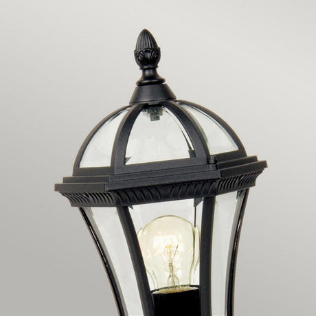 The Ledbury 1 Light Pedestal Lantern - Black is an outdoor lantern crafted from cast aluminum, featuring a classic black finish and a clear glass enclosure that showcases an exposed bulb. Its top is adorned with a decorative finial, set against a soft, neutral gray background for added elegance in your exterior lighting decor.