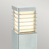 The Halmstad Large Bollard - Silver is a modern outdoor lamp post made from galvanized steel, showcasing a rectangular shape with a grid-like metal frame. This fixture casts a soft, warm glow against a plain gray background.