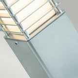 The close-up image highlights the Halmstad Small Bollard in Silver, featuring a modern design with a rectangular galvanized steel base and a frosted glass top. Horizontal metal bars wrap around the glass to elegantly diffuse light. Rated IP65, it is built to endure various weather conditions, against a plain light gray background.