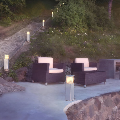 An outdoor seating area features two wicker chairs with white cushions on a stone patio. Halmstad Small Bollard - Silver lighting, boasting an IP65 rating, lines the pathway up a small hill surrounded by lush greenery and grass, ensuring durability and charm in any weather.