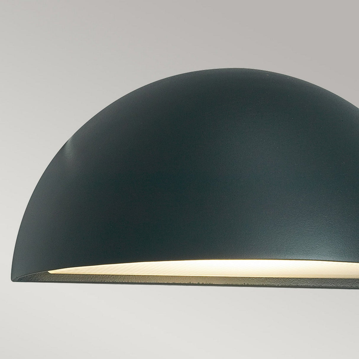 Close-up of the Halden Outdoor 1 Light Down Wall Light in graphite, featuring a sleek, dome-shaped design in dark green that casts a soft, warm glow. The lamp stands out gracefully against a neutral, light gray background.