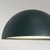 Close-up of the Halden Outdoor 1 Light Down Wall Light in graphite, featuring a sleek, dome-shaped design in dark green that casts a soft, warm glow. The lamp stands out gracefully against a neutral, light gray background.