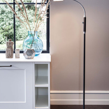 Vegas LED Floor Lamp 7W in black, shown beside a window with a slim black column, chrome gooseneck arm and flat head, placed near a white cabinet with decorative vases.