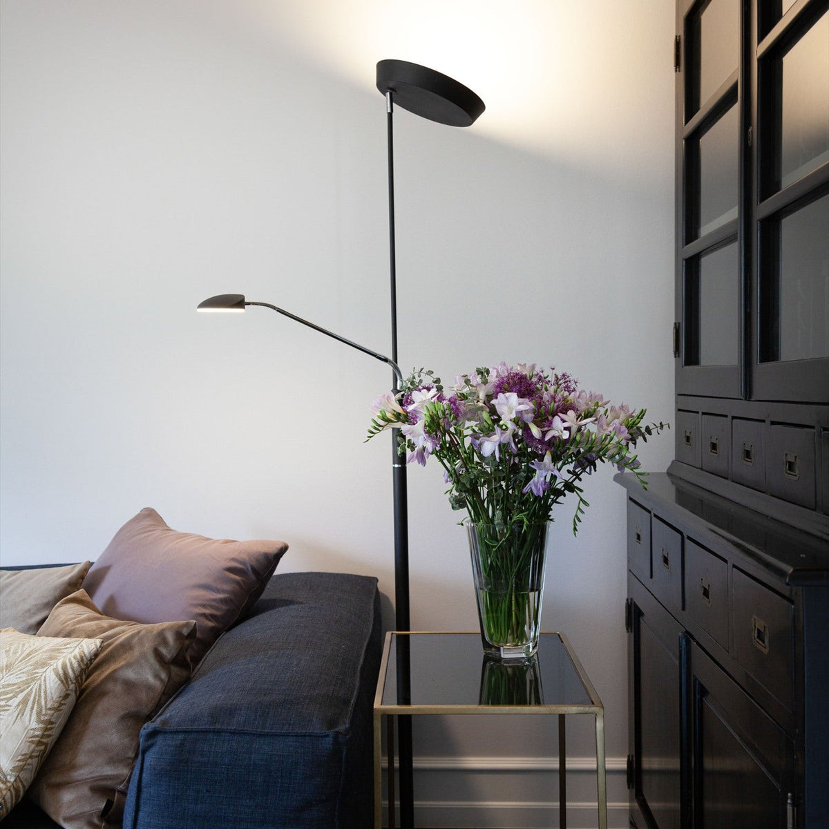 Vegas LED Combi Floor Lamp 20W in black, with a slim column, round base, upward-facing uplighter dish, and adjustable reading head, shown beside a sofa in a living room setting.