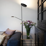 Vegas LED Combi Floor Lamp 20W in black, with a slim column, round base, upward-facing uplighter dish, and adjustable reading head, shown beside a sofa in a living room setting.