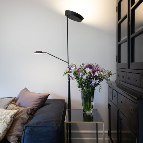Vegas LED Combi Floor Lamp 20W in black, with a slim column, round base, upward-facing uplighter dish, and adjustable reading head, shown beside a sofa in a living room setting.