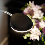 Vegas LED Combi Floor Lamp 20W in black, close-up of the oval reading head on a flexible chrome gooseneck arm, glowing edge visible against a blurred floral backdrop.