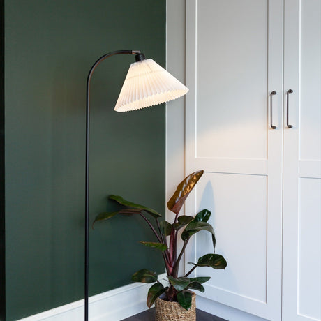 Medina Floor Lamp in black and white, with a pleated conical shade angled from a curved black stem, shown beside a plant against a green wall.