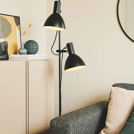 Baltimore 2 Light Floor Lamp in black, slim rail with two adjustable cone shades at different heights, cream interiors, shown beside a sofa in a living room.