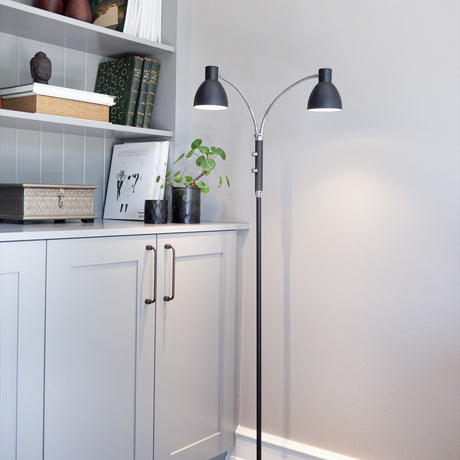 Hudson 2 Light Floor Lamp in black with slim column, chrome gooseneck arms and adjustable black cone heads, shown beside a grey shelving unit and cabinets.