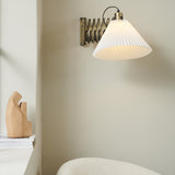 Medina X wall light in antique brass with an extendable scissor arm and wide pleated white fabric shade, shown illuminated on a neutral wall above a wooden decor piece.