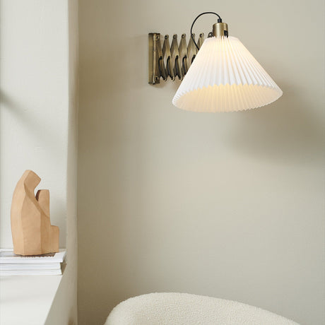 Medina X wall light in antique brass with an extendable scissor arm and wide pleated white fabric shade, shown illuminated on a neutral wall above a wooden decor piece.