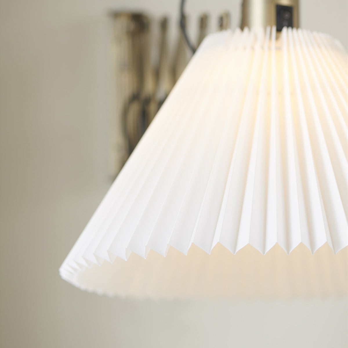 Medina X Wall Light in antique brass, close-up of the pleated white conical shade lit from within, with the lattice scissor arm visible behind against a neutral wall.