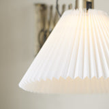 Medina X Wall Light in antique brass, close-up of the pleated white conical shade lit from within, with the lattice scissor arm visible behind against a neutral wall.