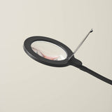 Magni LED X3 Dioptri Floor Lamp in black, close-up of the round magnifying lens head with lit border and hinged cover, mounted on a slim curved stem, shown against a plain light background.