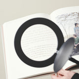 Magni LED X3 Dioptri Floor Lamp in black, shown in use magnifying open book text through its round lens head with lit border, held over pages by hand.