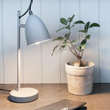 Yep Table Lamp in light grey with tapered dome shade, chrome pivot joint, and round base, shown on a desk beside a potted plant.