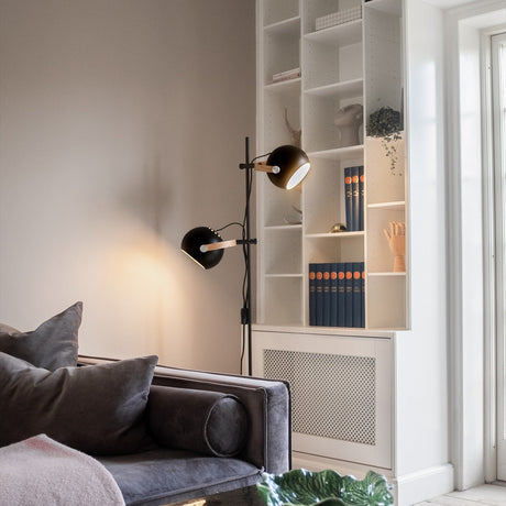 DC 2 Light Floor Lamp in black and oak, with a slim stem and two adjustable black spheres on oak arms, shown beside a bookcase in a living room.