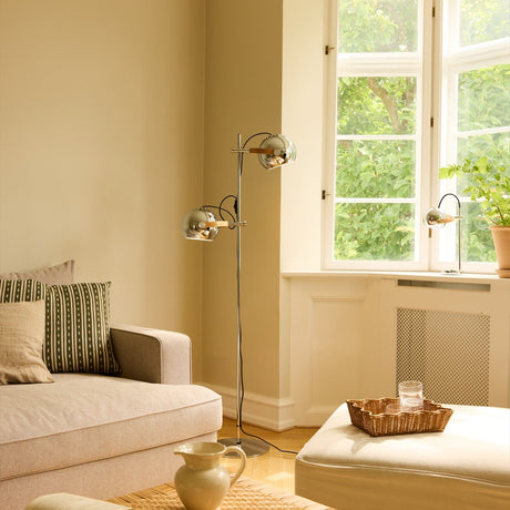 DC 2 Light Floor Lamp in chrome with two adjustable sphere shades on oak arms, shown in a living room beside a window.