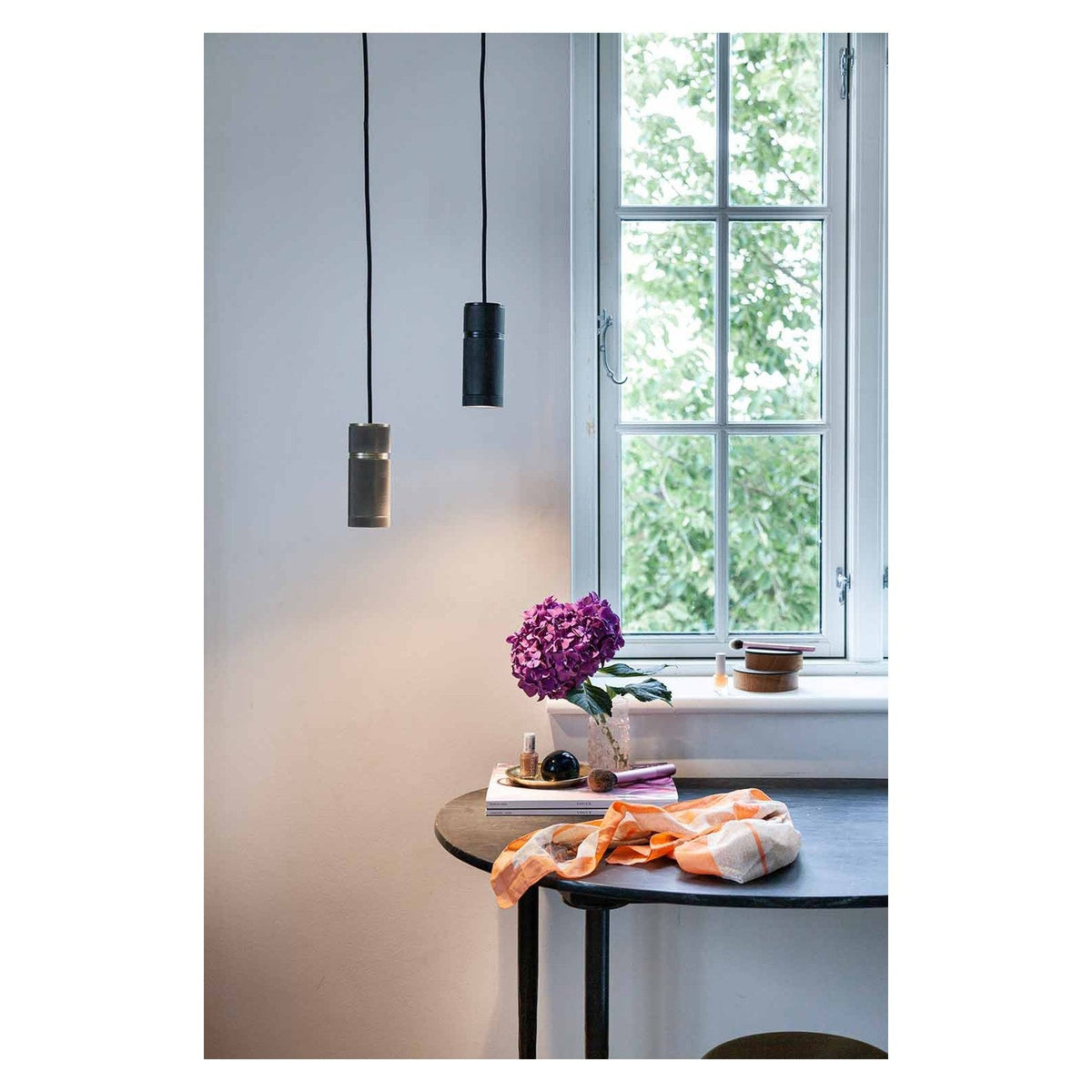 Halo Small Pendant Light in Black, a compact cylindrical fixture with knurled matt black finish, shown hanging above a dining table near a window.