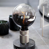 Halo Table Lamp in gunmetal and marble, knurled cylindrical metal base on a polished black marble disc, fitted with a large smoke-tinted glass globe bulb showing a visible filament.