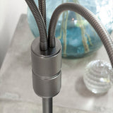 Close-up of the Halo 3 Light Floor Lamp's gunmetal knurled joint where three flexible black arms merge, set against a blurred room with a glass vase and decor.