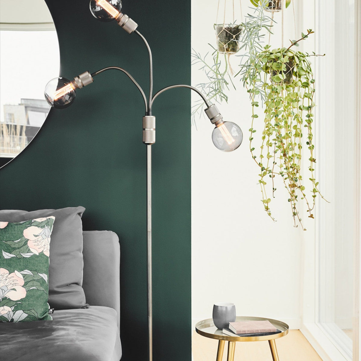Halo 3 Light Floor Lamp in gunmetal, with slim stem and three curved arms holding smoked glass globes, shown beside a dark green wall and hanging plants.