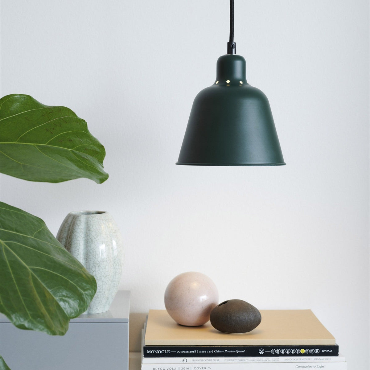 Carpenter Pendant Light 15cm in dark green with cone-shaped metal shade and vent holes, hanging above a shelf with books, a vase and a plant in a living room setting.