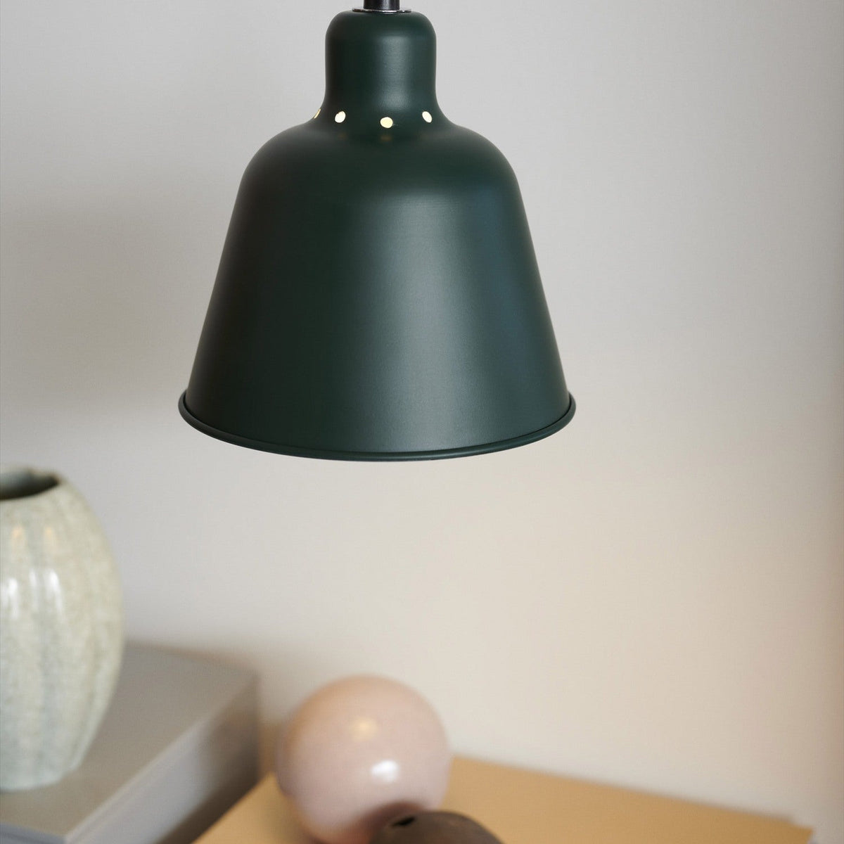 Carpenter Pendant Light 15cm in dark green, bell-shaped metal shade with vent holes on shoulder, shown hanging in a room setting above a shelf with decor.