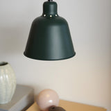 Carpenter Pendant Light 15cm in dark green, bell-shaped metal shade with vent holes on shoulder, shown hanging in a room setting above a shelf with decor.