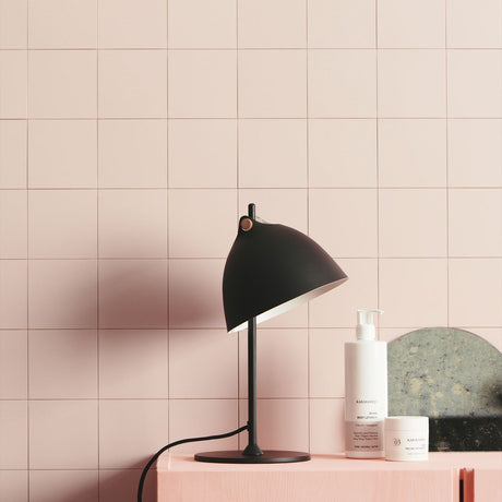 Aarhus Table Lamp 18cm in black and wood, angled dome shade with cream interior on a slim black post and round base, shown on a pink cabinet against pink tiled wall.