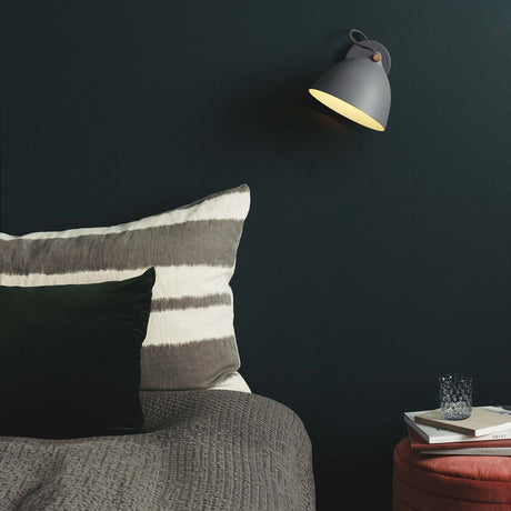 Aarhus Wall Light 18cm in grey with wood accent, dome shade angled downward on a wall plate, mounted above a bed with striped cushions in a dark-walled bedroom.