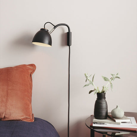 Nivå Level Wall Light in matte black with a bell-shaped shade and flexible arm, shown lit above a bed with rust cushion and navy throw beside a bedside table.