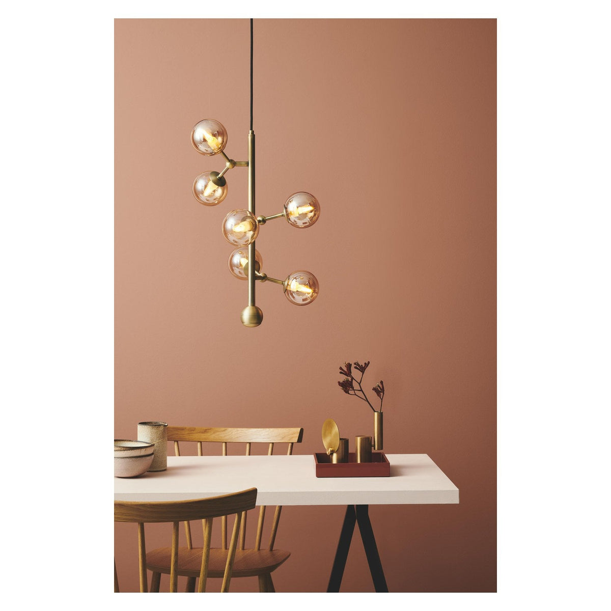 Atom Vertical 6 Light Chandelier in antique brass with amber glass spheres branching up a vertical rod, hung above a dining table in a warm terracotta room.