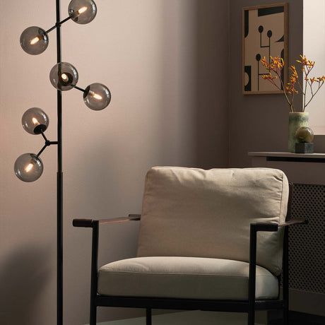 Atom Glass 6 Light Floor Lamp in black and smoke, with smoked glass spheres branching from a black rod on a round base, shown lit beside an armchair.