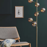 Atom 6 Light Floor Lamp in antique brass with amber glass spheres branching up the rod, shown beside a grey armchair and framed print in a living room.
