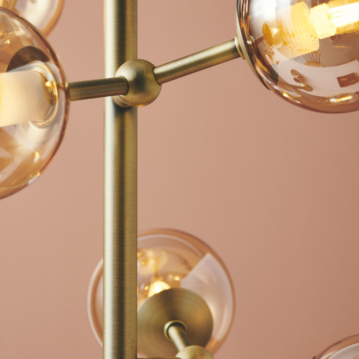 Close-up of the Atom 6 Light Floor Lamp's antique brass rod and joints, with amber glass globes housing visible bulbs against a soft pink background.