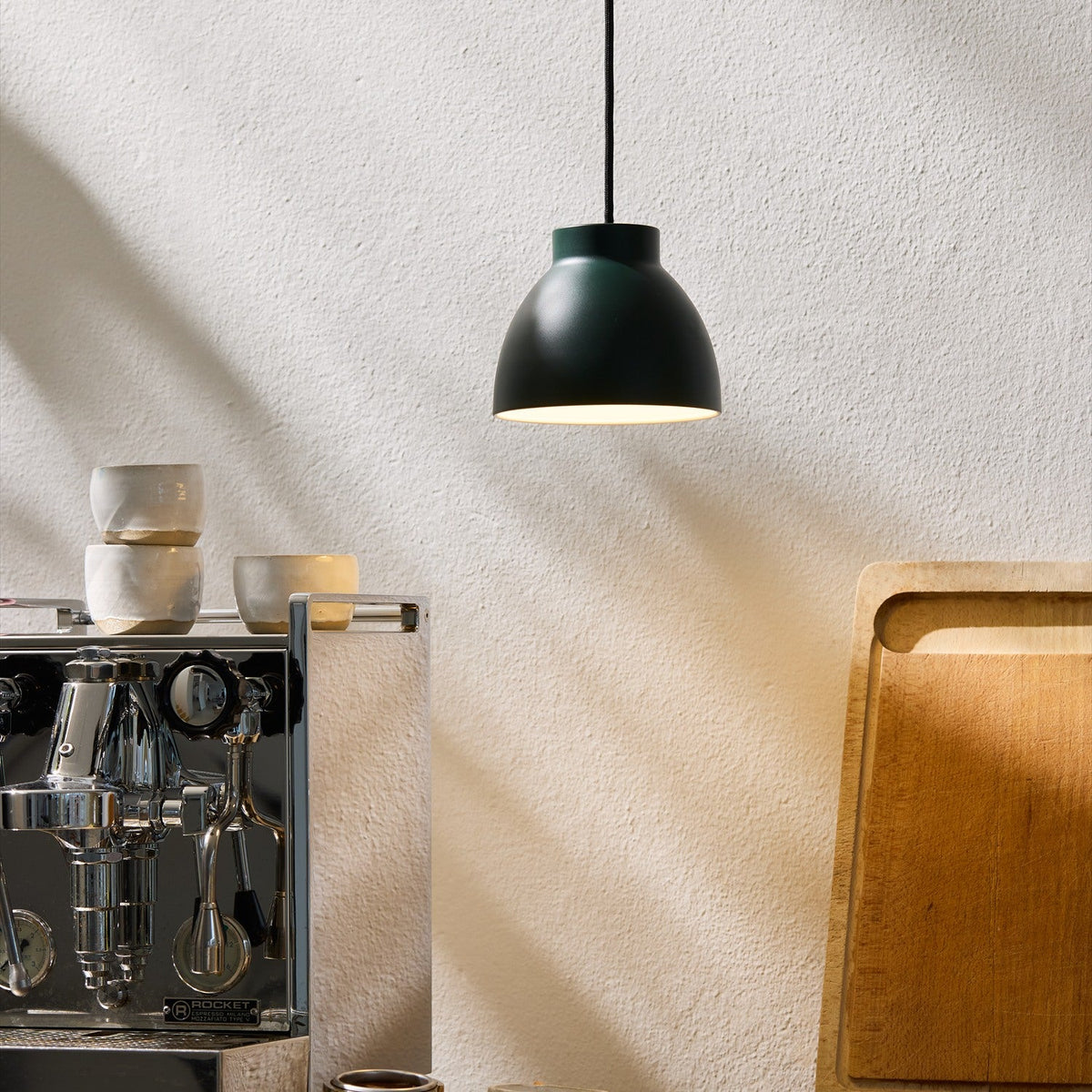 Object Pendant Light 16cm in green, a dome-shaped shade with matt chalky exterior and white interior, shown hanging above a kitchen counter with espresso machine and chopping board.