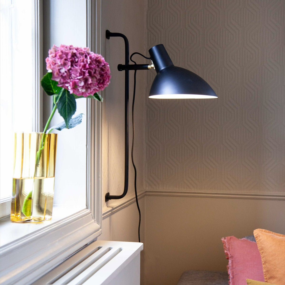 Metropole Grande Wall Light in black, wide angled dome shade with brass collar detail on a wall-mounted rail, shown lit beside a window in a living room.