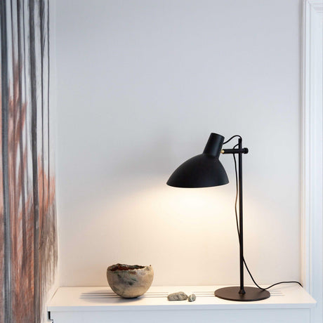 Metropole Table Lamp in black, with a wide matt black dome shade on a slim upright and round base, shown lit on a white shelf beside a stone bowl and pebbles.