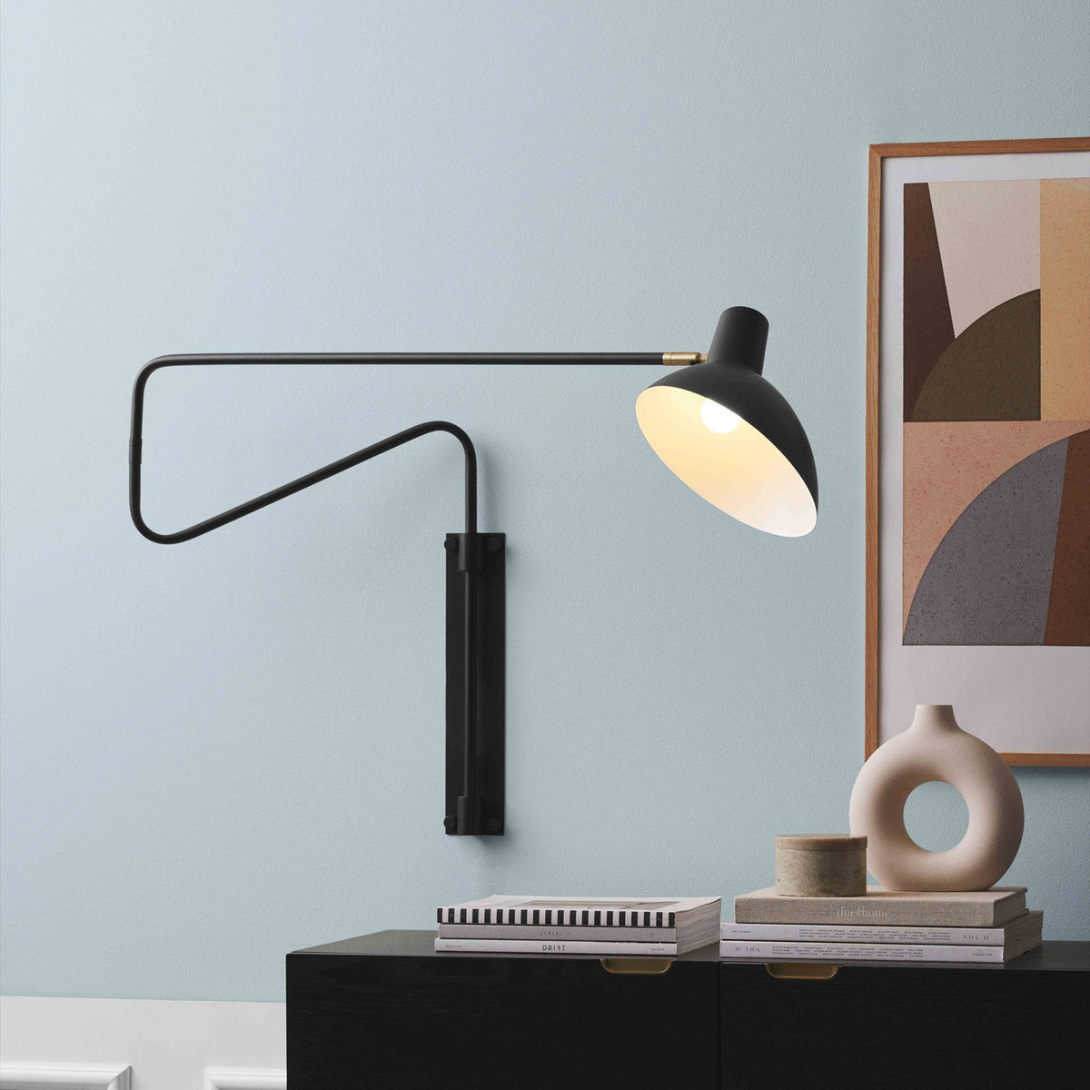 Metropole Deluxe Swing Wall Light in black, with a wide shallow dome shade on a jointed folding arm and brass knuckle detail, shown above a dark sideboard against a pale blue wall.