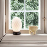 Twist Oval Table Lamp in opal and black, egg-shaped twisted glass shade on a black half-dome base, lit on a windowsill beside a small ceramic bowl.