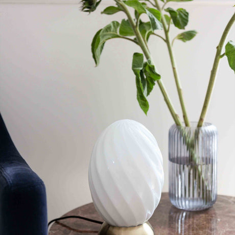 Twist Oval Table Lamp in opal glass with a twisted flute pattern and antique brass base, styled on a side table beside a ribbed glass vase with foliage.