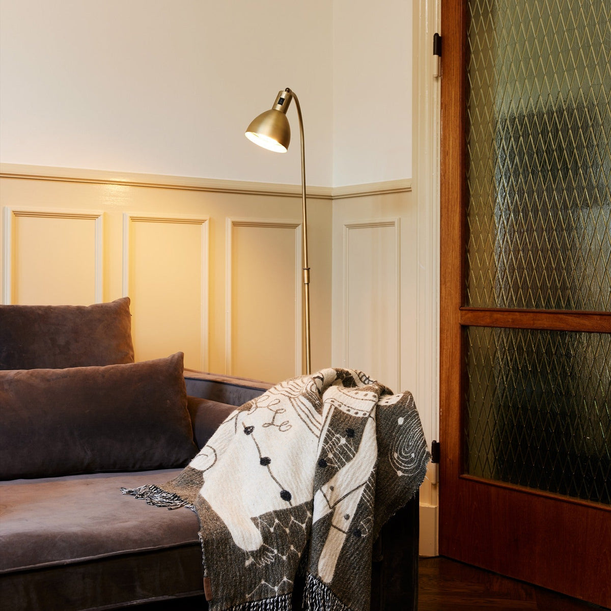 Angora Floor Lamp in antique brass with curved stem and angled dome shade, shown lit beside a sofa with a patterned throw in a living room.