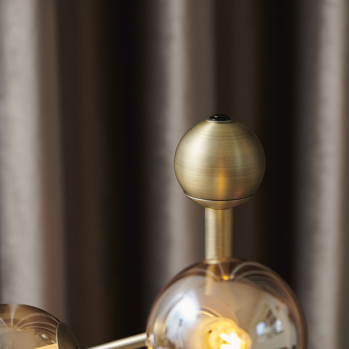 Close-up of the Atom Glass 4 Light Table Lamp's antique brass stem and finial ball, with an amber glass globe and lit G9 bulb visible below.