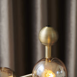 Close-up of the Atom Glass 4 Light Table Lamp's antique brass stem and finial ball, with an amber glass globe and lit G9 bulb visible below.